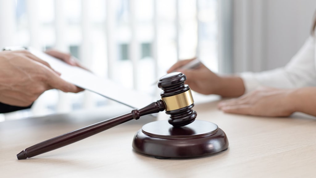 A wooden judge's gavel on a desk between two people, one handing over a document and the other holding a pen, suggesting a legal or official agreement.